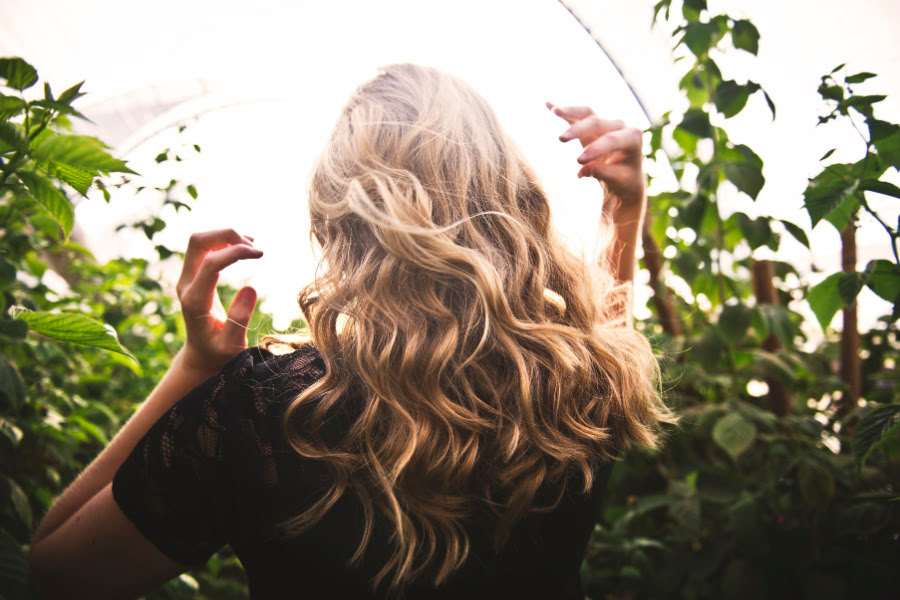 Blonde Frau mit langen welligen Haaren steht von hinten inmitten grüner Pflanzen im warmen Sonnenlicht.