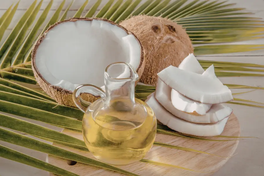 Kokosnussöl in einer Glasflasche mit halbierter Kokosnuss, frischen Kokosstücken und grünen Palmblättern auf einem hellen Holzbrett.