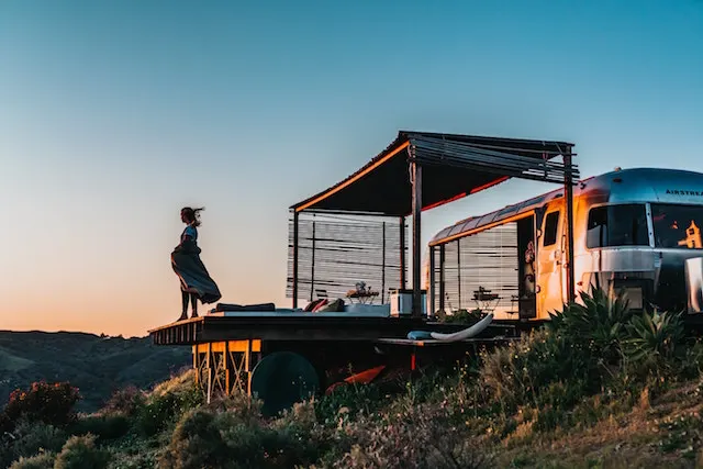 Frau auf einer Holzterrasse vor einem silbernen Airstream-Wohnwagen bei Sonnenuntergang in den Bergen - camping