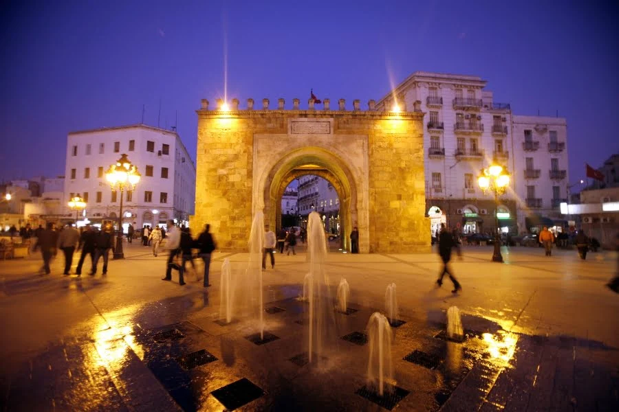 Tunesien entdecken: Aktuelle Trends, Sehenswürdigkeiten und kulinarische Highlights 17 Ihr seht ein beleuchtetes Stadttor in Tunis bei Abenddämmerung mit einem Brunnen im Vordergrund und Menschen, die über den Platz laufen.
