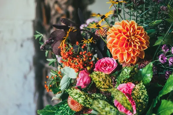 Ein bunter Blumenstrauß mit orangefarbener Dahlie, rosa Rosen, Beerenzweigen und gemischtem Blattwerk vor unscharfem Hintergrund