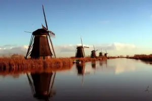 Mehrere traditionelle Windmühlen entlang eines ruhigen Kanals in den Niederlanden, gespiegelt im Wasser unter blauem Himmel.