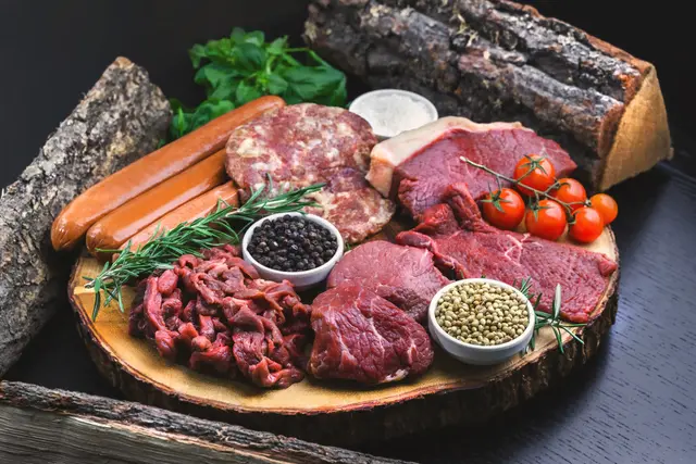 Es ist eine große Holzscheibe zu sehen. Auf der Holzscheibe liegen unterschiedliche Arten von rohem Fleisch, 3 Würstchen, ein Strauch Cocktailtomaten, Gewürze und Kräuter. Im Hintergrund liegen noch drei Holzscheite. 