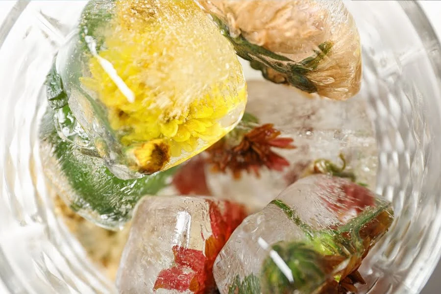 Klare Eiswürfel mit eingefrorenen Blüten, Kräutern und Früchten in einer Glasschale.