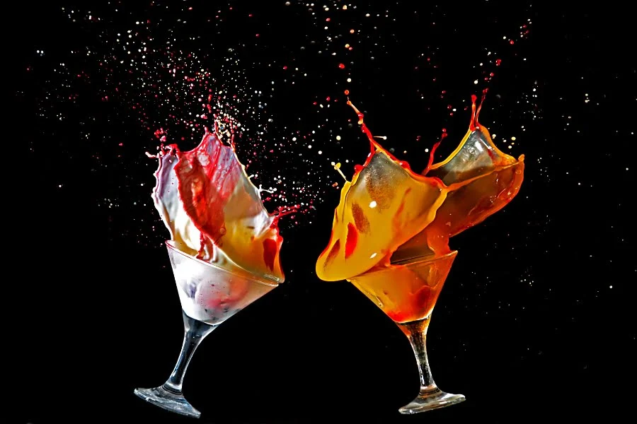 Farbige Cocktails spritzen aus zwei Cocktailgläsern vor schwarzem Hintergrund mit spektakulären Flüssigkeitseffekten.