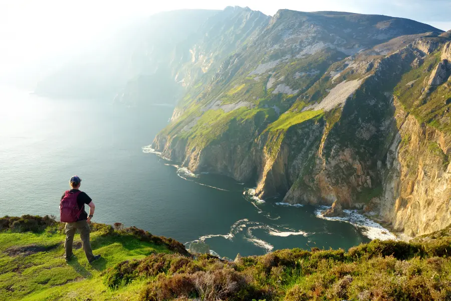 Sea-Trekking, CrossGolf & Co.: Diese Outdoor-Sportarten sind diesen Sommer ein Muss 3 Wanderer mit Rucksack blickt von einer grünen Klippe auf steile Küstenfelsen und das Meer