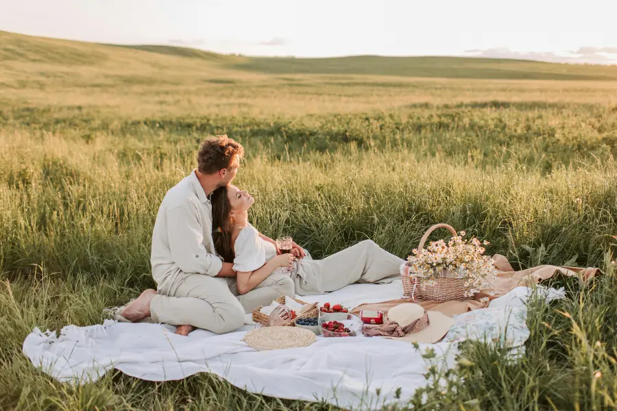 Paar sitzt entspannt auf einer Picknickdecke in einer grünen Wiese bei Sonnenuntergang mit Korb und Snacks