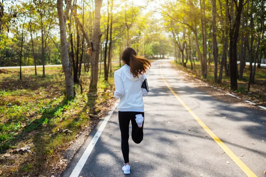 Jogging-Point Gutschein Person joggt auf einem asphaltierten Waldweg, umgeben von hohen Bäumen und warmem Sonnenlicht an einem ruhigen Tag.