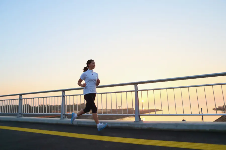 Jogging-Point Gutschein Läuferin joggt bei Sonnenaufgang oder Sonnenuntergang über eine Brücke am Wasser und genießt das Training im Freien.