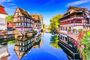 Historische Fachwerkhäuser am Fluss in Straßburg mit spiegelglatter Wasseroberfläche und blauem Himmel.