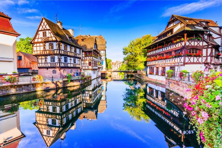 Historische Fachwerkhäuser am Fluss in Straßburg mit spiegelglatter Wasseroberfläche und blauem Himmel.
