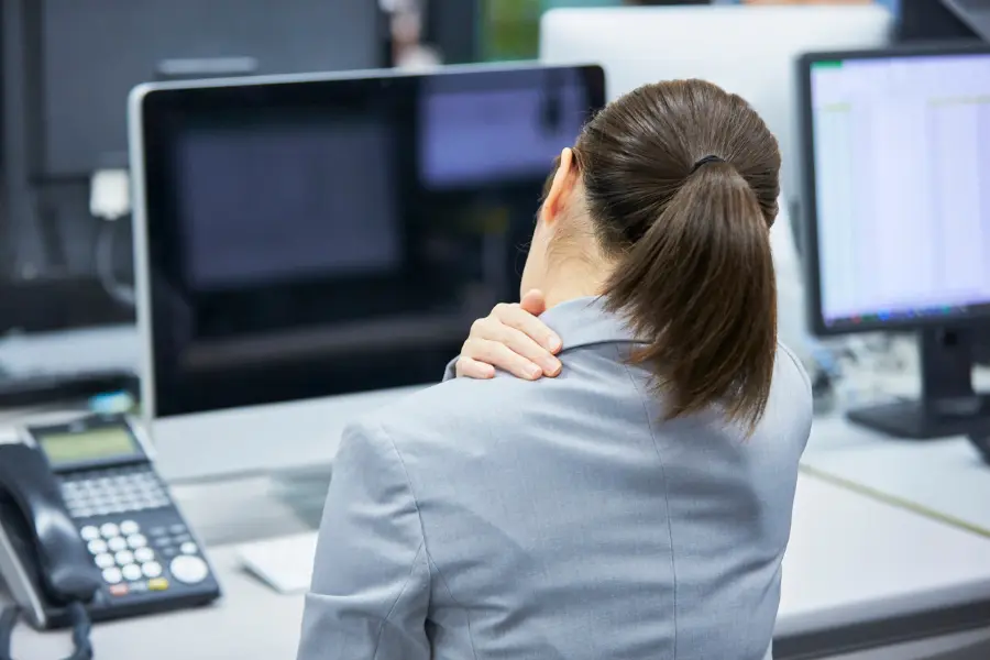 Eine Qual im Alltag: Was hilft gegen Rücken- und Gliederschmerzen? 1 Frau im Büro massiert ihren Nacken, während sie vor mehreren Computerbildschirmen sitzt – Symbolbild für Nackenschmerzen und Verspannungen am Arbeitsplatz.