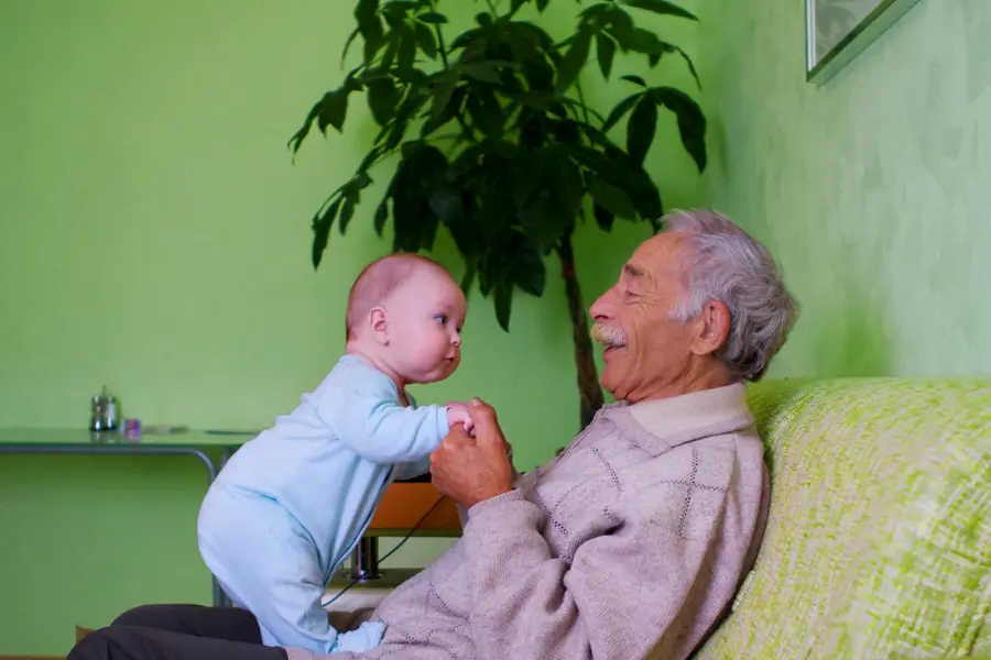 Tag der Senioren - was wir von unseren Großeltern lernen können 9 Lächelnder Großvater hält die Hände eines Babys, das vor ihm auf dem Sofa steht – ein liebevoller Moment zwischen zwei Generationen.