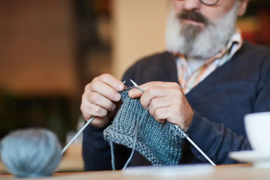 Tag der Senioren - was wir von unseren Großeltern lernen können 7 Älterer Mann mit grauem Bart sitzt am Tisch und strickt mit grauer Wolle – ein humorvolles Beispiel für Geschick, Geduld und kreative Fähigkeiten im Alter.