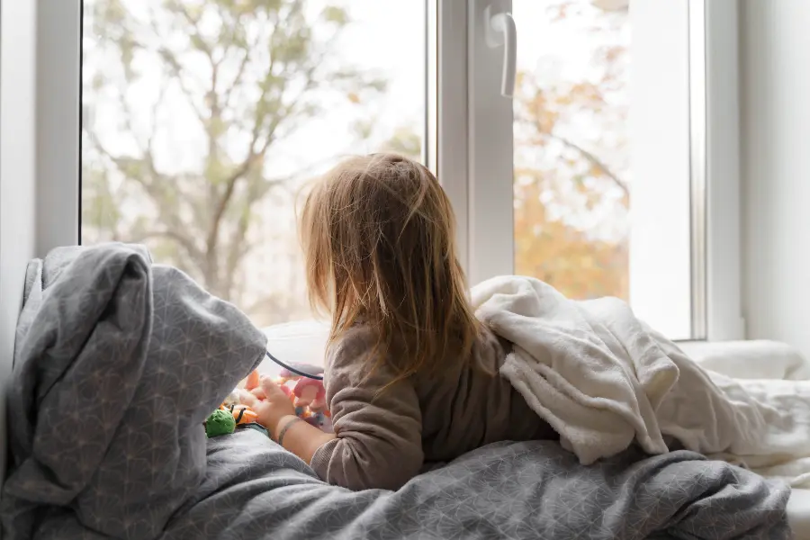 Rabatt Coupons Kind liegt mit einer weichen Decke im gemütlichen Bett am Fenster und blickt nach draußen auf die herbstliche Landschaft.