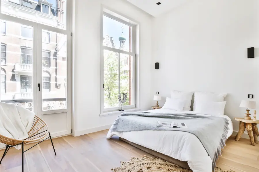 Helles Schlafzimmer mit großen Fenstern, weißen Wänden, Naturholzboden, weißem Bett und minimalistischem Dekor.