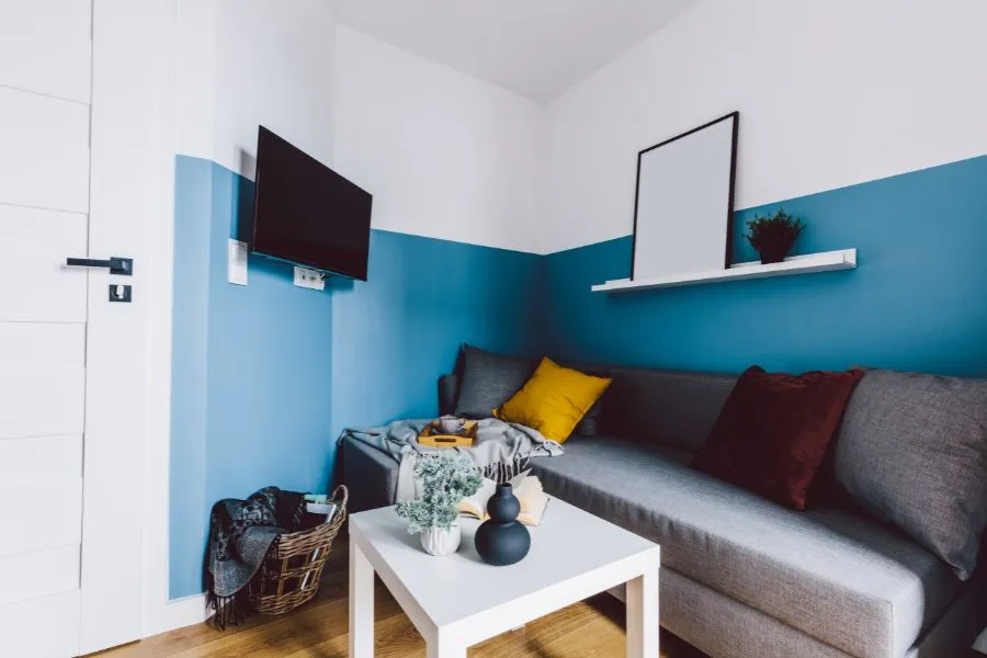 Kleines Wohnzimmer mit blauer Wand, grauem Sofa, kleinem Couchtisch, bunten Kissen und einem schmalen Wandregal unter einem Fernseher.