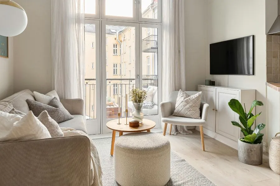 Helles, stilvoll eingerichtetes Wohnzimmer mit Sofa, Sessel, rundem Holztisch, Fernseher und bodentiefen Fenstern in einem kleinen Raum.