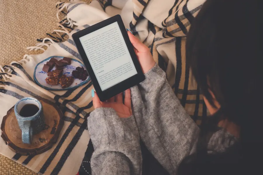 Person liest gemütlich auf einem E-Reader, eingekuschelt in eine Decke, mit Kaffee und Süßigkeiten daneben.