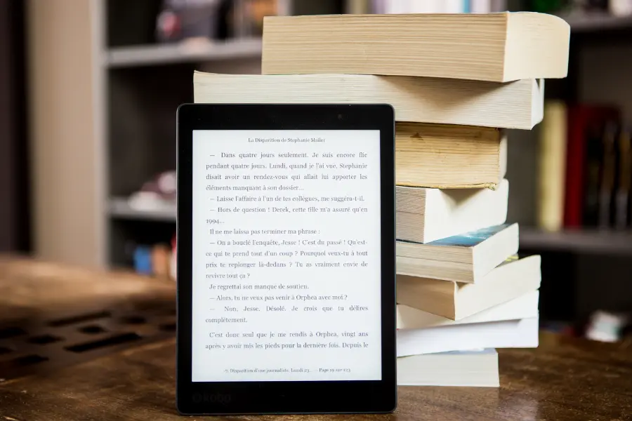 E-Reader mit geöffnetem Text steht vor einem Stapel gedruckter Bücher auf einem Tisch.