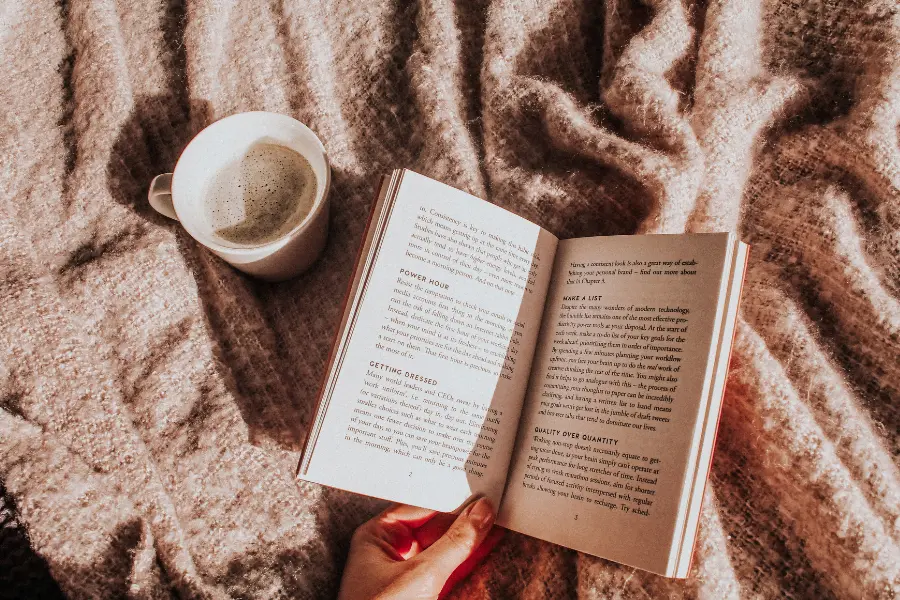 Geöffnetes Buch und Tasse Kaffee auf einer weichen Decke in gemütlicher Lesestimmung.