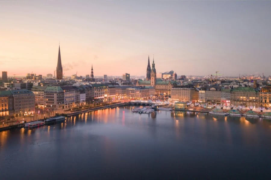 Abendlicher Blick auf Hamburg mit Binnenalster, beleuchteter Uferpromenade, historischen Gebäuden und Kirchtürmen im Hintergrund.