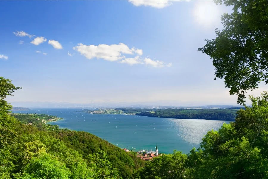 Blick über einen türkisblauen See mit Segelbooten, grünen Wäldern und sonnigem Himmel.