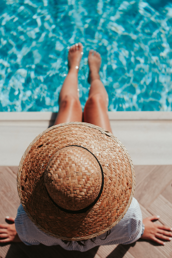 Person mit Strohhut sitzt am Poolrand und lässt die Füße ins Wasser hängen.