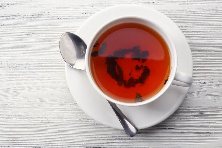 Teesorten und ihre Wirkung - diese Fakten habt ihr garantiert noch nicht gekannt | www.rabatt-coupon.com Tasse mit frisch aufgebrühtem schwarzem Tee und sichtbaren losen Teeblättern auf weißem Holztisch.