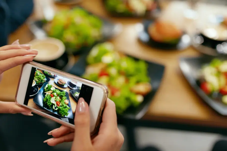 Person fotografiert mit einem Smartphone mehrere Salate und Speisen auf einem gedeckten Tisch.