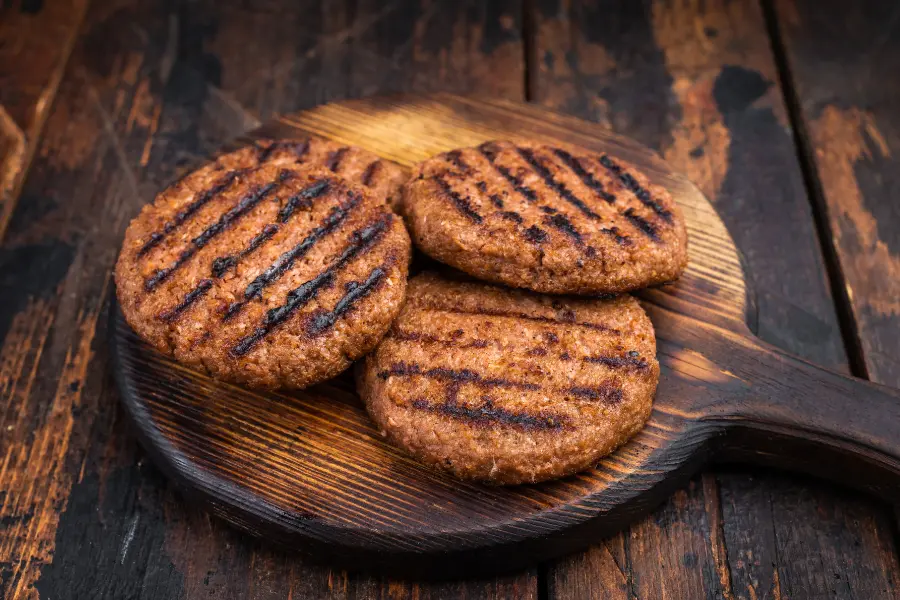 Drei gegrillte Burger-Patties mit Grillstreifen liegen auf einem rustikalen Holzbrett.