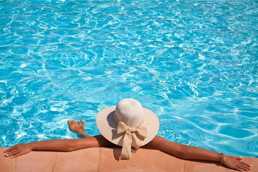 Frau mit großem Strohhut lehnt entspannt am Rand eines Pools mit ausgebreiteten Armen im Wasser.