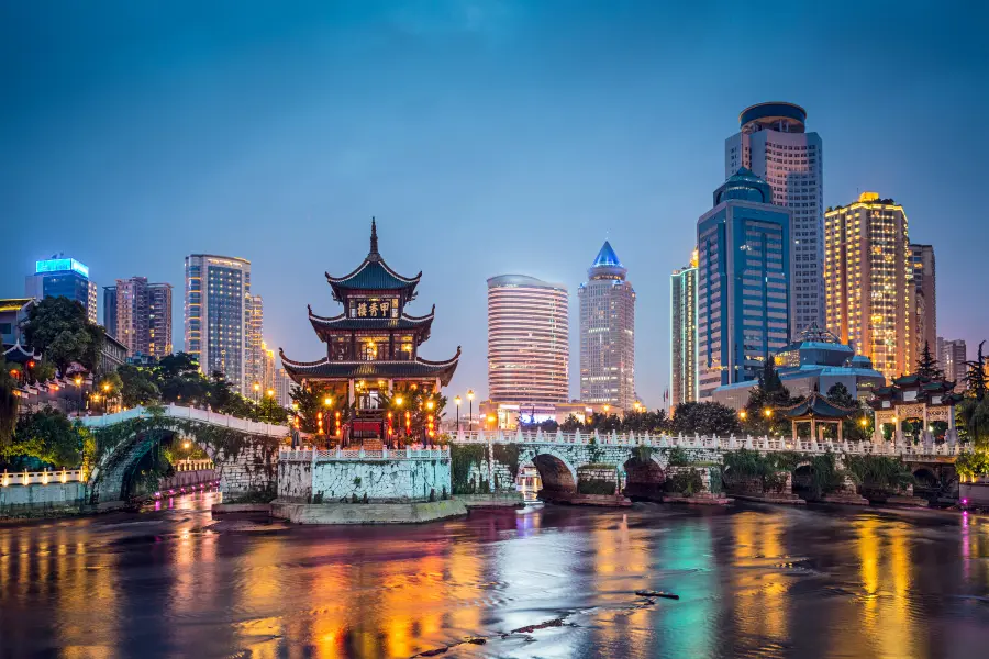 Illuminated city skyline at dusk with traditional Chinese architecture, modern skyscrapers, a river, and reflections on the water.