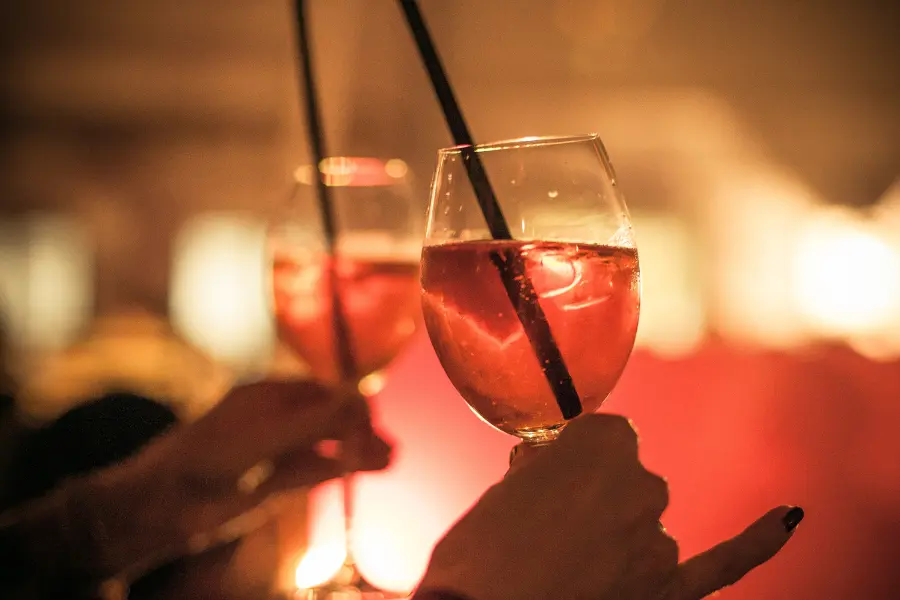 Unvergessliche Aktivitäten mit Freunden könnt ihr euch mit dem Wassersport Europa Gutschein sichern. Two people holding glasses of pink cocktails with straws in a warm, softly lit bar or lounge setting.