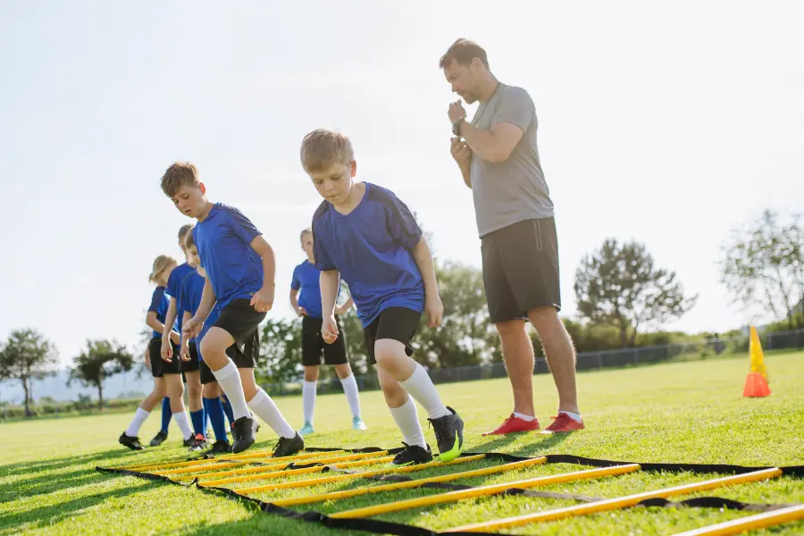 Ihr spielt im Jugendfußball und Corona hat euch nicht die Motivation am spielen genommen? Dann könnt ihr mit dem Sportlädchen Gutschein tolles Equipment zum Sparpreis ergattern. Young soccer players in blue uniforms practicing footwork drills on an agility ladder while a coach supervises on a sunny sports field.