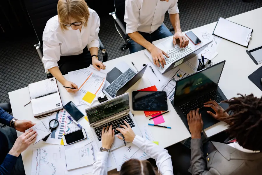 Sichert euch mit dem Legimi Gutschein Rabatte auf Hörbücher zum Thema Arbeitnehmerrecht in der Ausbildung mit Schulabschluss Team of professionals working together around a shared office table with laptops, documents, charts, notebooks, and digital devices.
