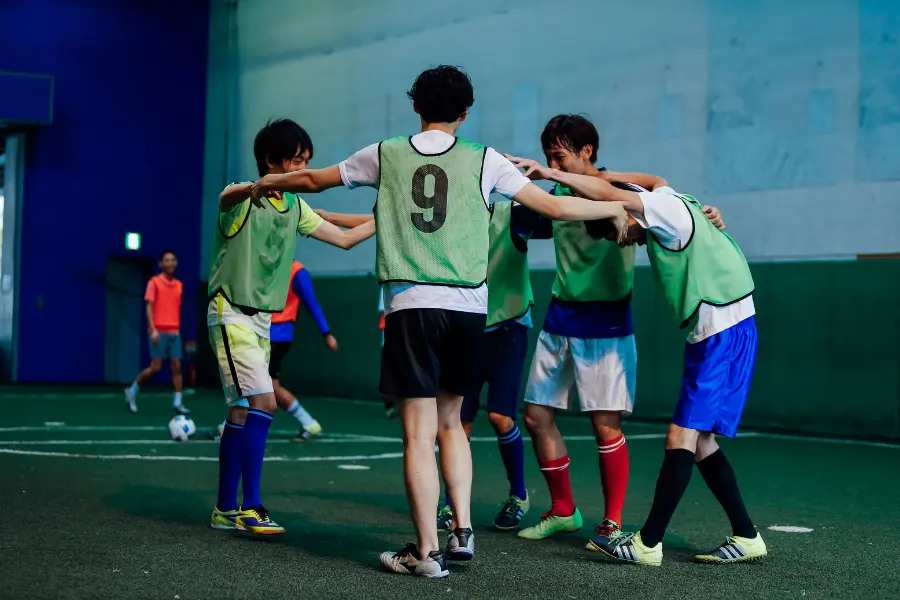 Ihr spielt Jugendfußball und Corona hat euch aus dem Schwung gebracht? Mit dem Sportlädchen Gutschein findest du genau das richtige Equipment und an dir zu arbeiten und kannst zugleich von vielen Rabatten profitieren. Indoor soccer players wearing green training bibs huddle together with arms around each other during a team practice session.