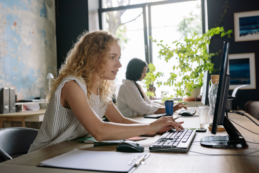 Mit der Office Discount Aktion, können sich selbst Azubis Büro-Artikel leisten. Two women working at desktop computers in a modern office with natural light, plants, and shared workspace desks.
