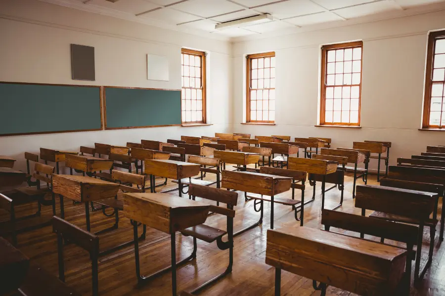 Empty classroom with rows of wooden desks, chalkboards, and tall windows in a traditional school setting.