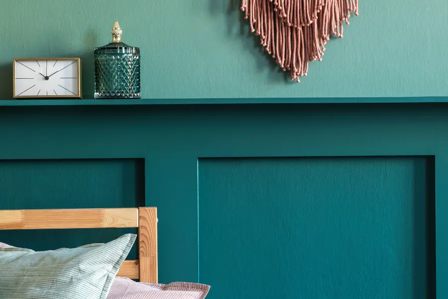 Teal bedroom wall with wooden bed frame, pillows, shelf decor, a small clock, glass jar, and woven wall hanging.