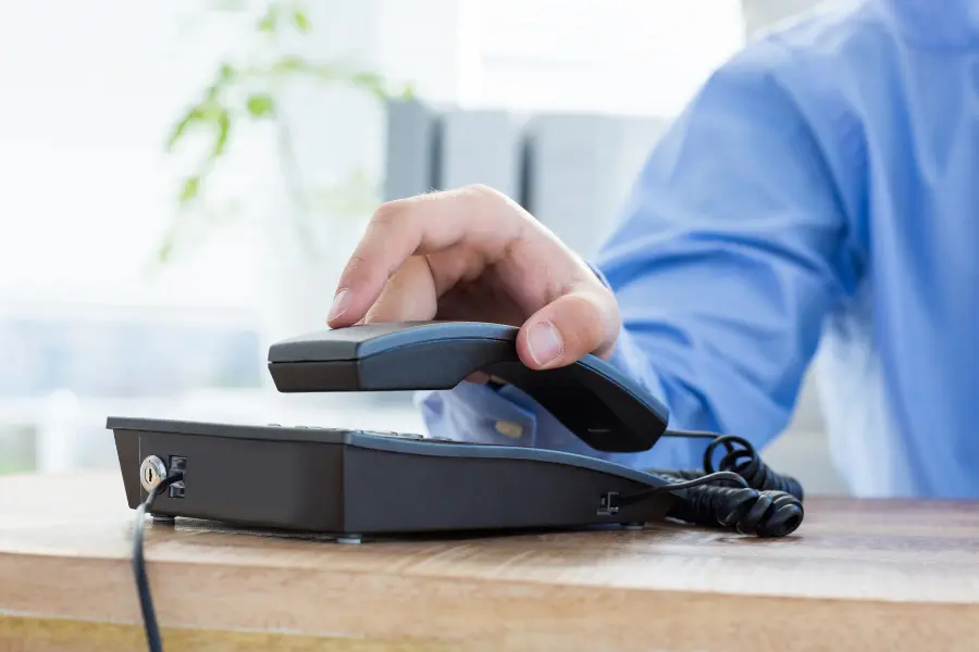 Hand lifting the receiver of a desk phone in a bright office setting.