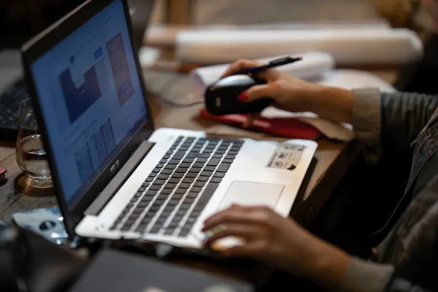 Person working on a laptop and using a computer mouse at a desk in a dimly lit workspace.