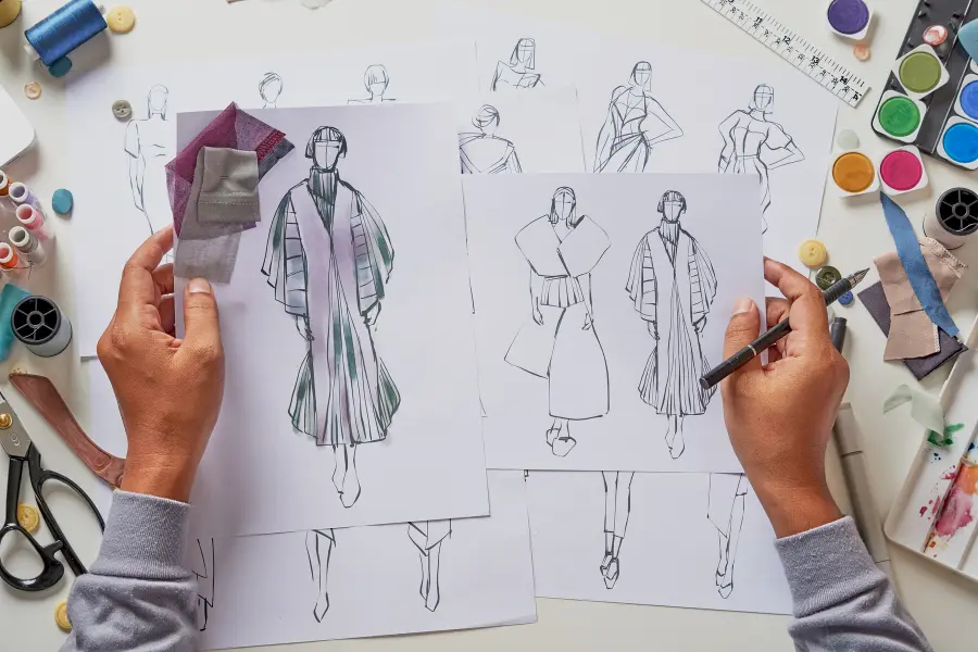 Jetzt euer Moodboard vervollständigen mit den Artikeln von Napapijri Gutschein. Schaut vorbei und sichert euch immer die neusten Angebote. Fashion designer working on clothing sketches with fabric swatches, art supplies, scissors, and design tools spread across a creative