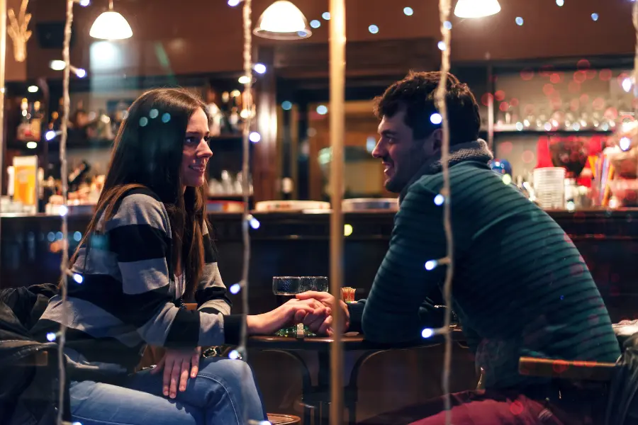 Couple smiling and holding hands during a romantic date in a warmly lit restaurant with decorative string lights.
