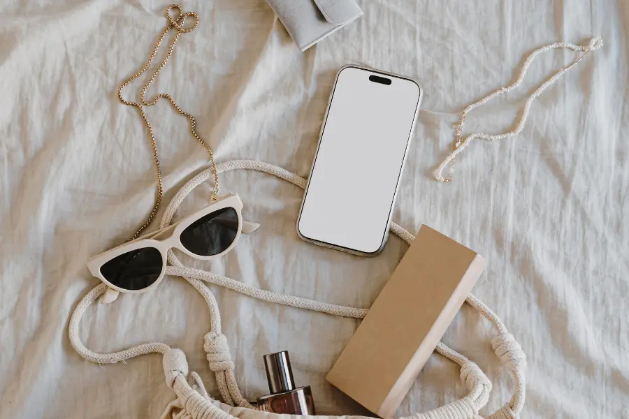 Jetzt euer Outfit kreativer gestalten mit den Taschen von "der offizielle Taschen Shop". Spart auf eure Lieblingstaschen mit den neusten Angeboten. Smartphone with blank screen placed on a white bed beside sunglasses, jewelry, perfume, a book, and a woven bag.
