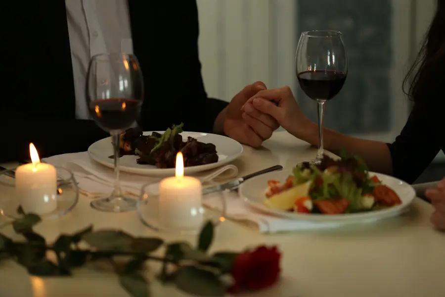 Couple holding hands during a romantic candlelit dinner with wine glasses, plated food, and a red rose on the table.