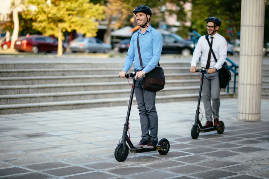 Sichert euch mit dem Jacob Elektronik Gutschein Rabatte zugunsten der Elektromobilität in Deutschland Two business commuters wearing helmets ride electric scooters through an urban plaza.