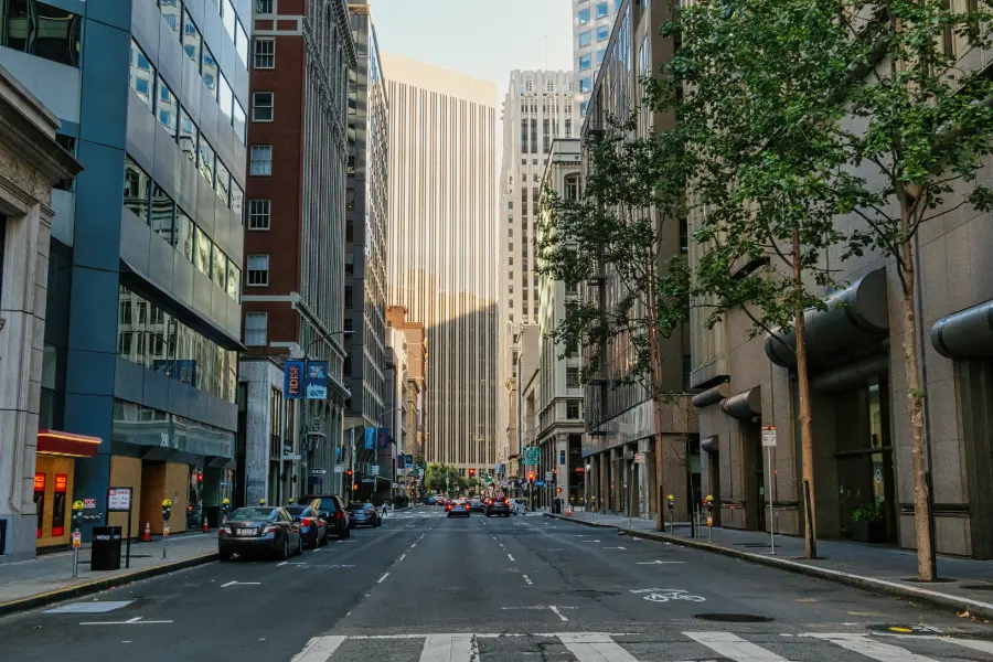 Urban downtown street lined with tall office buildings, trees, parked cars, and traffic in the city center.