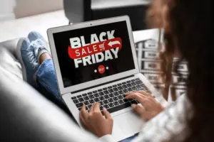 Eine Person sitzt auf einem Sofa und nutzt ein Laptop. Auf dem Bildschirm ist ein großes „Black Friday“-Motiv zu sehen, bestehend aus schwarzen und roten Schriftzügen sowie einem Einkaufswagensymbol. Die Hände der Person liegen auf der Tastatur. Der Raum ist hell und modern eingerichtet.
