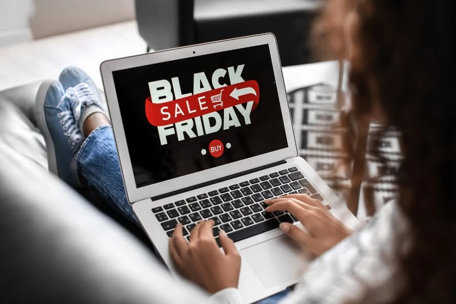 Eine Person sitzt auf einem Sofa und nutzt ein Laptop. Auf dem Bildschirm ist ein großes „Black Friday“-Motiv zu sehen, bestehend aus schwarzen und roten Schriftzügen sowie einem Einkaufswagensymbol. Die Hände der Person liegen auf der Tastatur. Der Raum ist hell und modern eingerichtet.